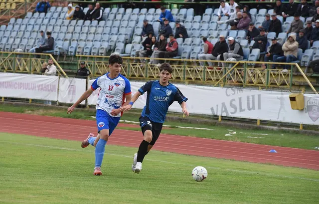 Doi jucători de fotbal de pe un teren de gazon verde aleargă spre un balon alb.