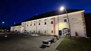 O clădire lungă, iluminată noaptea, cu ferestre mici și un spațiu pavat în față care conține câteva bănci de beton.