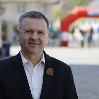 Un bărbat cu părul scurt și cămașă albă poartă un sacou negru cu un pin pe rever, zâmbind în prim-plan.