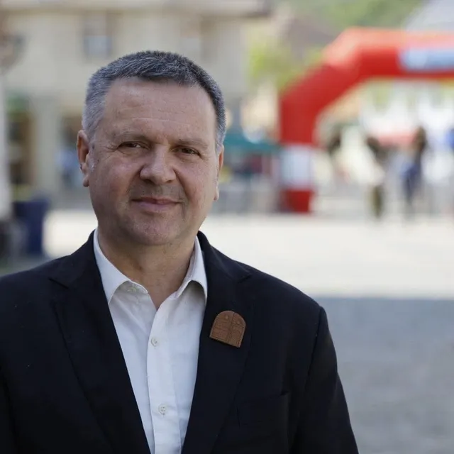 Un bărbat cu părul scurt și cămașă albă poartă un sacou negru cu un pin pe rever, zâmbind în prim-plan.