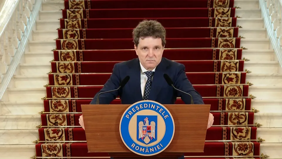 Un bărbat cu o cămașă albă, cravată și sacou stă în spatele unui podium de lemn cu emblema României, în fața unor scări cu covor roșu.