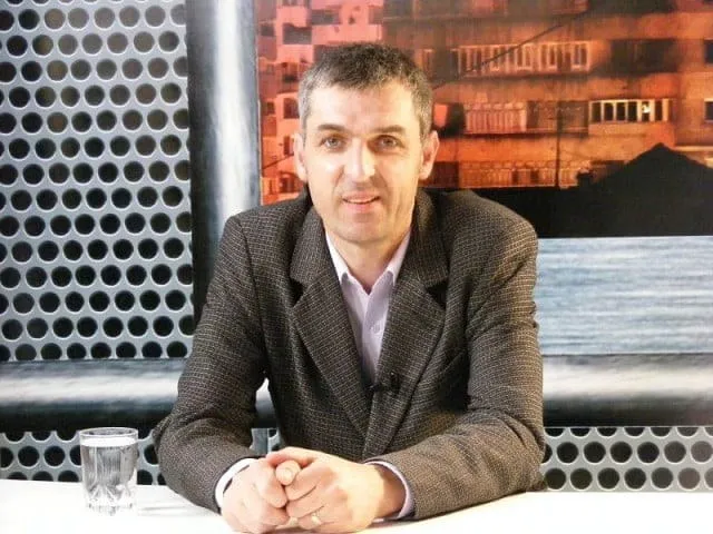 Un bărbat cu părul scurt și sacou maro stă la o masă albă lângă un pahar cu apă, în fața unui fundal cu textură și o imagine.