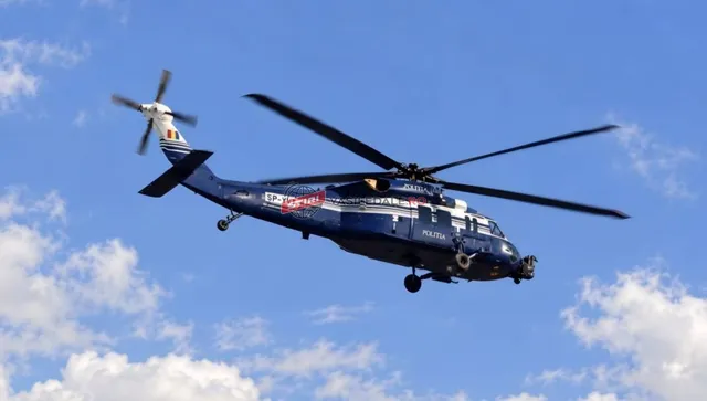 Un elicopter albastru cu marcaje albe și roșii zboară pe un cer albastru cu nori albi.