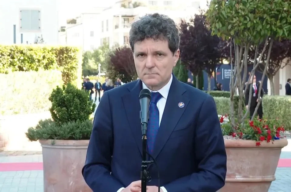 Un bărbat cu părul închis la culoare, purtând un costum albastru și cravată albastră, stă în fața unui microfon într-un spațiu exterior cu plante și clădiri.