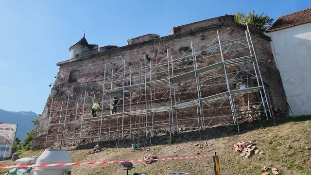 O structură de metal pentru construcții este montată pe un zid de cărămidă și piatră, pe o suprafață de deal în lumină naturală.
