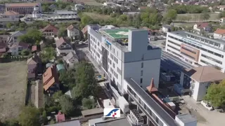 O vedere aeriană prezintă clădiri moderne de culoare albă și gri, înconjurate de case individuale cu acoperișuri înclinate și vegetație dispersă.