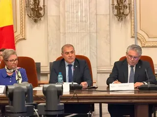 Trei persoane în costume oficiale stau la o masă lungă de conferințe într-o încăpere cu pereți de marmură și aplicații de iluminat clasice.