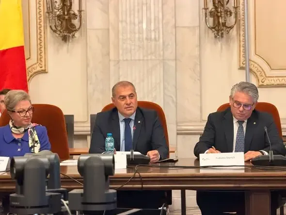 Trei persoane în costume oficiale stau la o masă lungă de conferințe într-o încăpere cu pereți de marmură și aplicații de iluminat clasice.