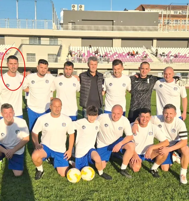 Un grup de bărbați îmbrăcați în echipamente sportive albe și blu sunt poziționați pe un teren de fotbal verde în fața unor tribune.