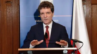 Un bărbat cu părul închis la culoare, purtând sacou închis și cravată roșie, stă în spatele unui podium în fața unui fundal albastru și a unui steag.
