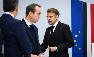 Trei bărbați în costume închise stau în picioare în fața unui fundal alb cu dungi albastre și roșii.