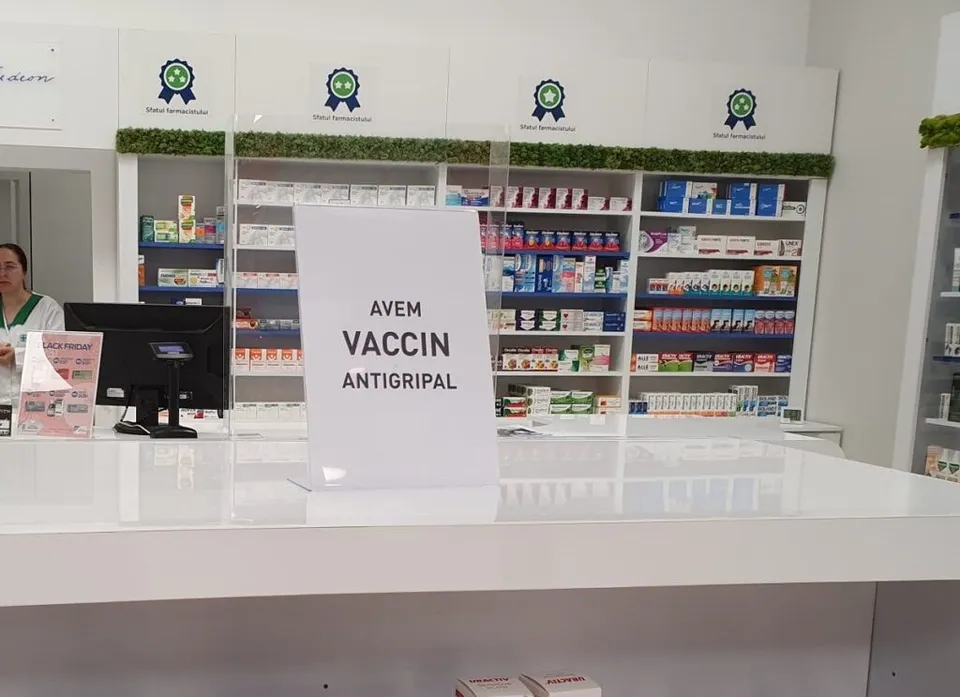 Un semn alb cu text negru stă pe un blat de farmacie lângă rafturi pline cu diverse cutii de medicamente colorate.