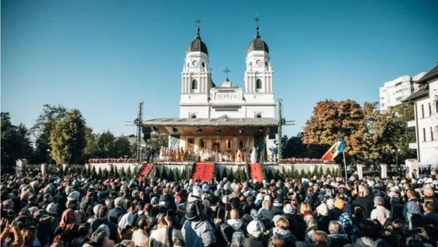 O mulțime de oameni stă în fața unei catedrale albe cu două turnuri, în timp ce un podiu este situat în prim-plan.