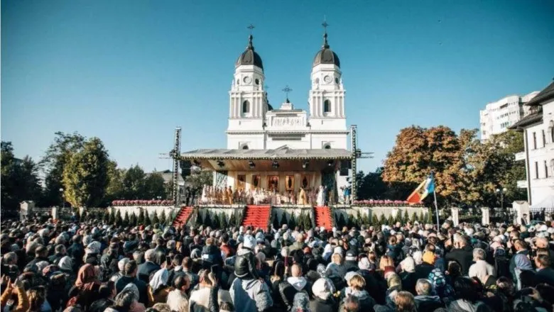 O mulțime de oameni stă în fața unei catedrale albe cu două turnuri, în timp ce un podiu este situat în prim-plan.