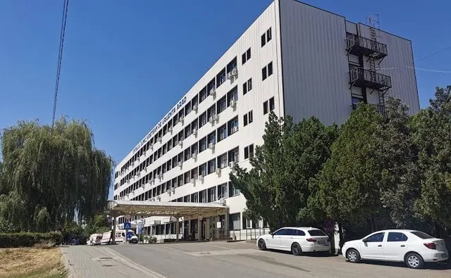 O clădire albă de mari dimensiuni cu ferestre multiple este situată lângă un drum unde sunt parcate două mașini albe.