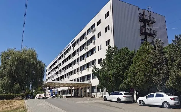 O clădire albă de mari dimensiuni cu ferestre multiple este situată lângă un drum unde sunt parcate două mașini albe.