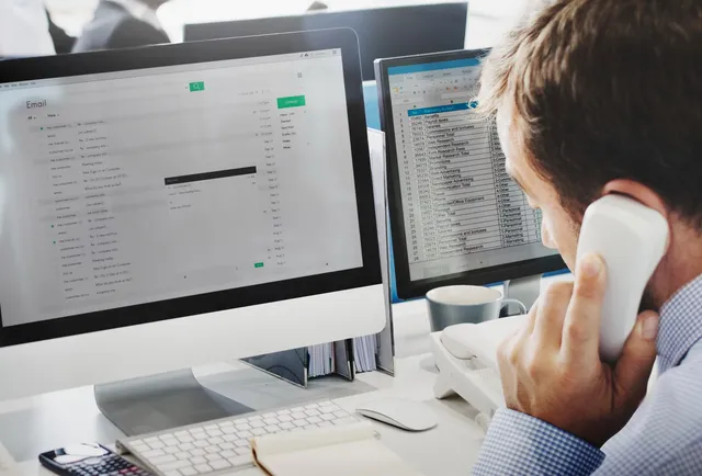 Un bărbat cu o cămașă albastră vorbește la telefon în timp ce privește două monitoare de calculator afișând interfețe de e-mail.