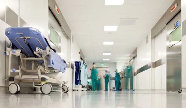 Un brancard albastru este poziționat pe un hol de spital cu pereți albi și persoane purtând uniforme medicale verzi în fundal.