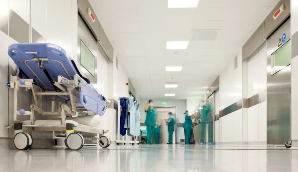 Un brancard albastru este poziționat pe un hol de spital cu pereți albi și persoane purtând uniforme medicale verzi în fundal.