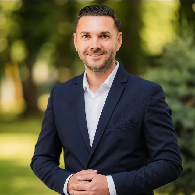 Un bărbat cu părul scurt și pielea deschisă poartă un sacou închis la culoare peste o cămașă albă, zâmbind în fața unui fundal verde.