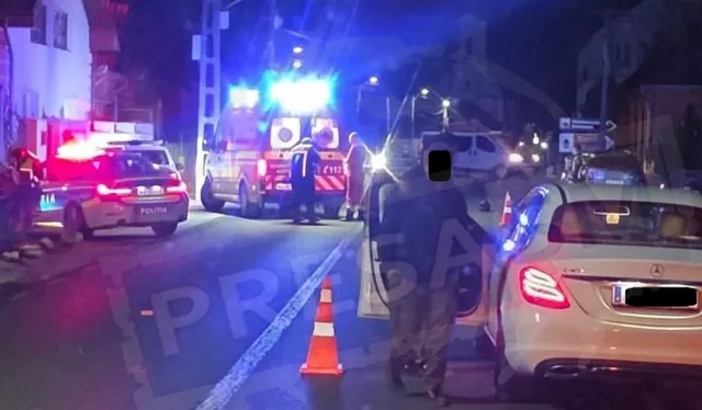 Vehiculele de poliție și o ambulanță cu luminile aprinse sunt parcate pe un drum de noapte lângă un autoturism alb.