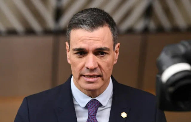 Un bărbat cu părul scurt și gri, purtând sacou închis la culoare, cămașă albastră și cravată mov, privește spre camera aflată în prim-plan.