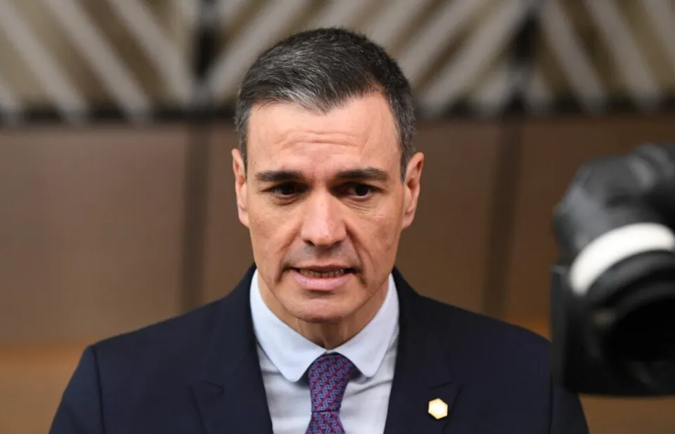 Un bărbat cu părul scurt și gri, purtând sacou închis la culoare, cămașă albastră și cravată mov, privește spre camera aflată în prim-plan.