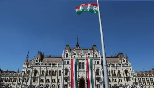 O steag tricoloragru de culoare roșu, alb și verde flutură pe un stârp înalt în fața unei clădiri monumentale de stil gotic sub un cer senin.
