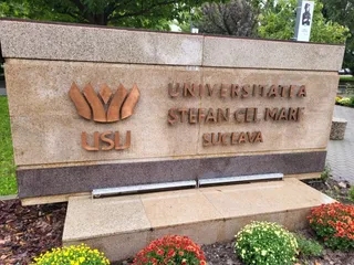 Un monument de piatră cu inscripția UNIVERＳITATEA ȘTEFAN CEL MARI SUCEAVA și un simbol metalic este situat lângă plante verzi și flori colorate.