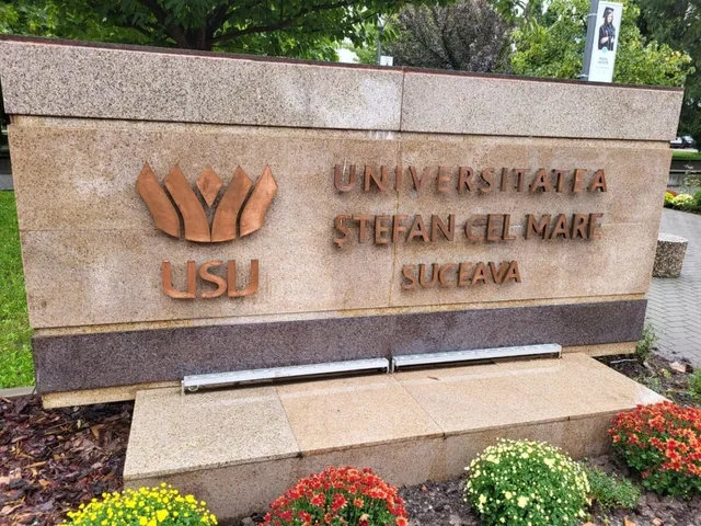 Un monument de piatră cu inscripția UNIVERＳITATEA ȘTEFAN CEL MARI SUCEAVA și un simbol metalic este situat lângă plante verzi și flori colorate.