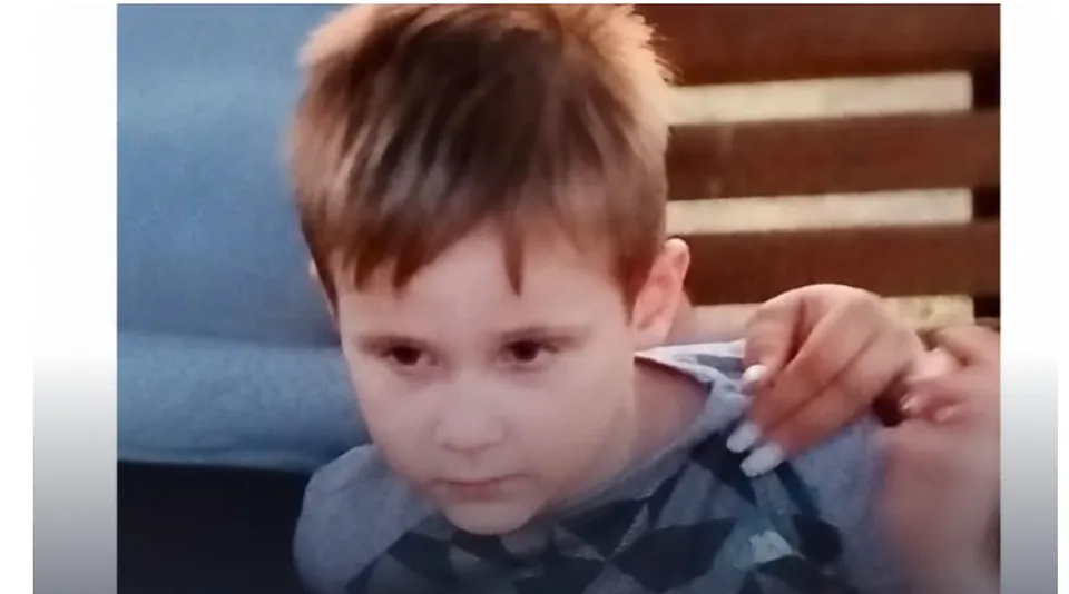 Un copil cu părul scurt și deschis la culoare poartă un tricou gri cu modele geometrice în culori închise.