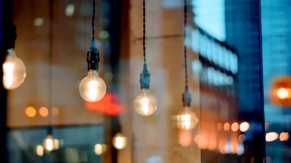 Mai multe becuri suspendate emit o lumină caldă într-un decor urban cu fundal desfocat.