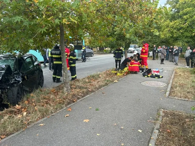 Pompieri în uniforme de lucru și persoane civile stau pe un trotuar lângă un autoturism negru avariat și un autobuz pe o stradă cu copaci.