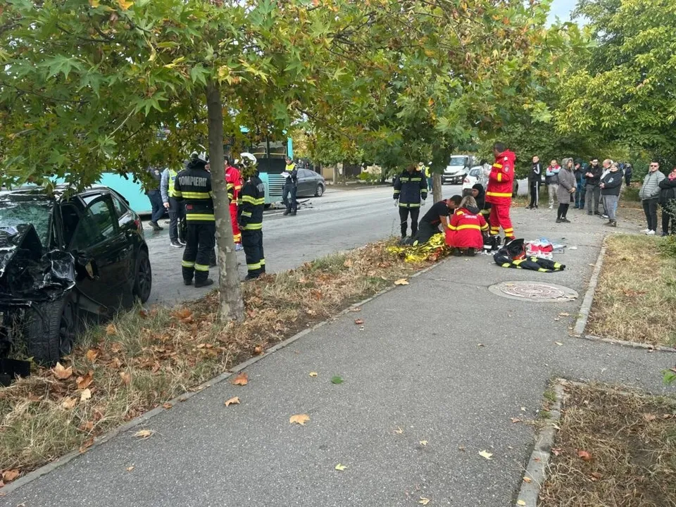 Pompieri în uniforme de lucru și persoane civile stau pe un trotuar lângă un autoturism negru avariat și un autobuz pe o stradă cu copaci.