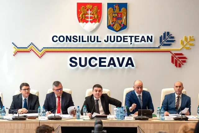 Cinci bărbați în costume purtând cravate stau așezați la o masă lungă în fața unui perete cu un logou și textul Consiliul Județean Suceava.