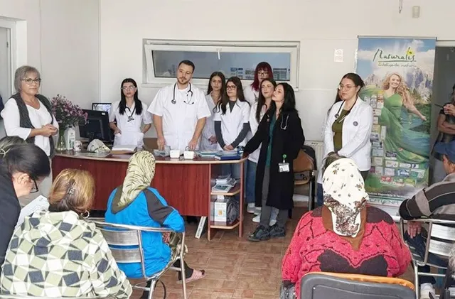 Un grup de persoane în halate albe stă în jurul unei mese într-o încăpere, în timp ce un grup de oameni stă pe scaune cu spatele la cameră.