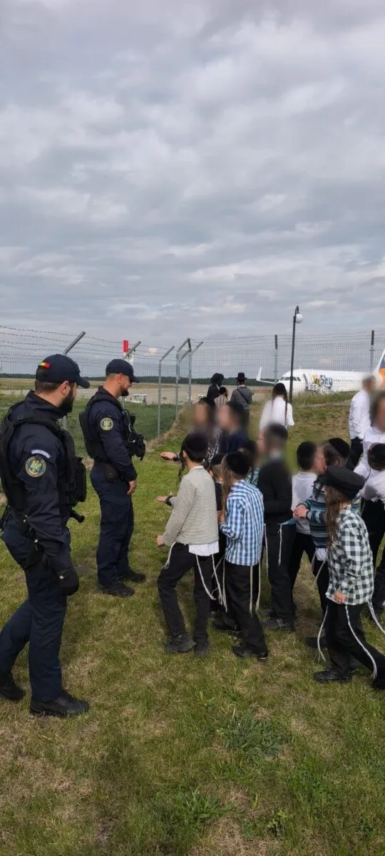 Un grup de persoane stă pe un câmp de iarbă lângă doi bărbați în uniforme închise și cu echipament de protecție.