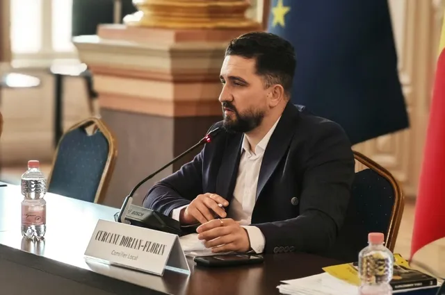 Un bărbat cu barbă, purtând sacou închis și cămașă albă, stă la o masă cu un microfon și o sticlă de apă în față.