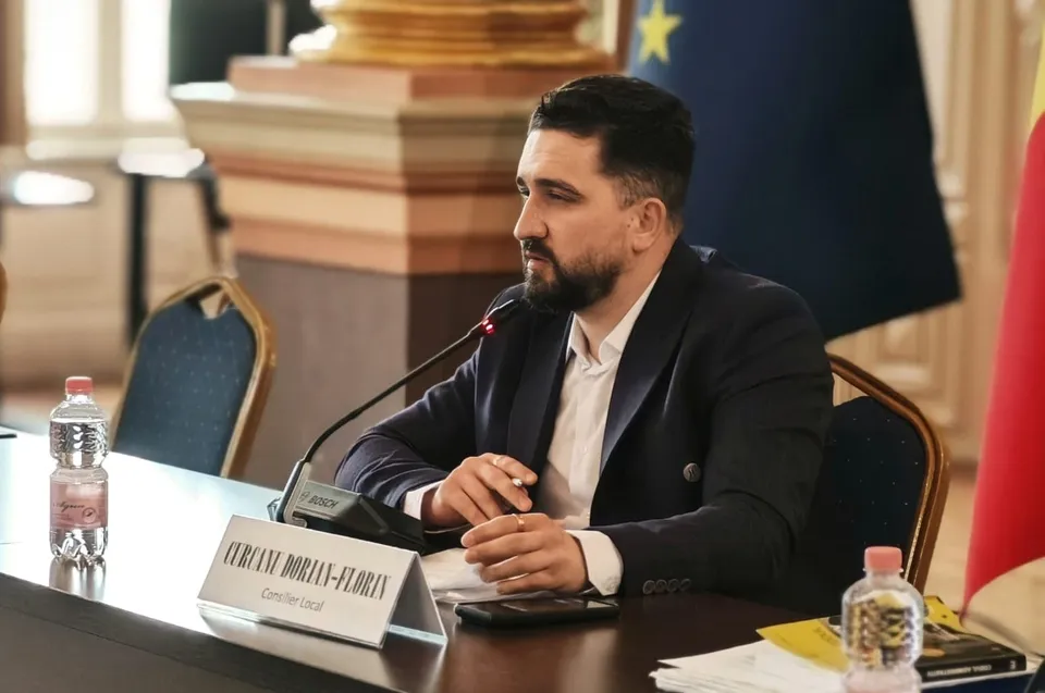 Un bărbat cu barbă, purtând sacou închis și cămașă albă, stă la o masă cu un microfon și o sticlă de apă în față.