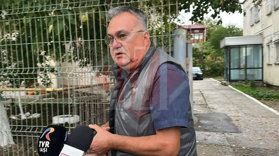 Un bărbat cu ochelari și vestă gri este microfonat de două microfoane în fața unui gard metalic și al unei clădiri.