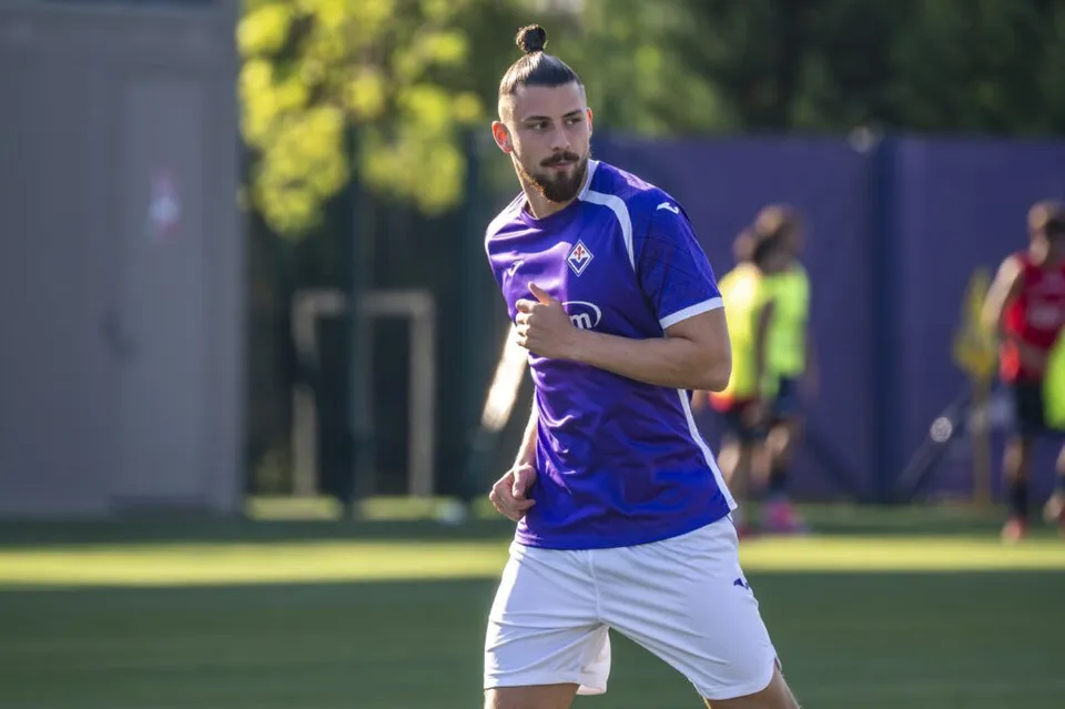 Un bărbat cu părul prins într-un coc și barbă poartă un tricou violet și pantaloni scurți albi pe un teren de fotbal verde.