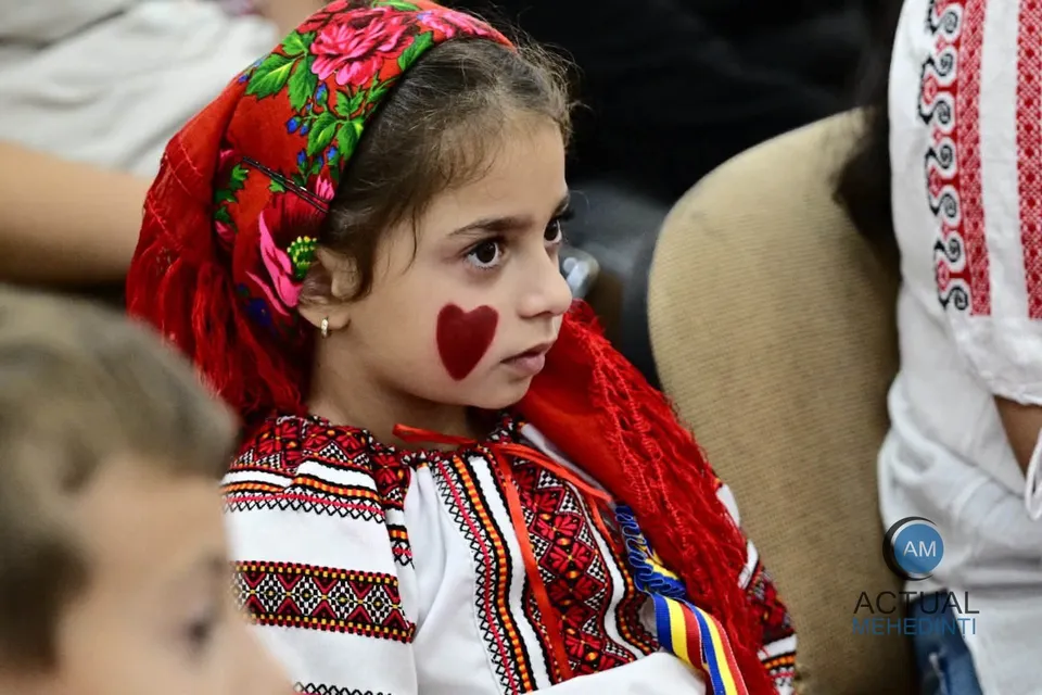 O copilă cu un desen în formă de inimă pe obraz poartă un basma roșie cu motive florale și o cămașă tradițională brodată.