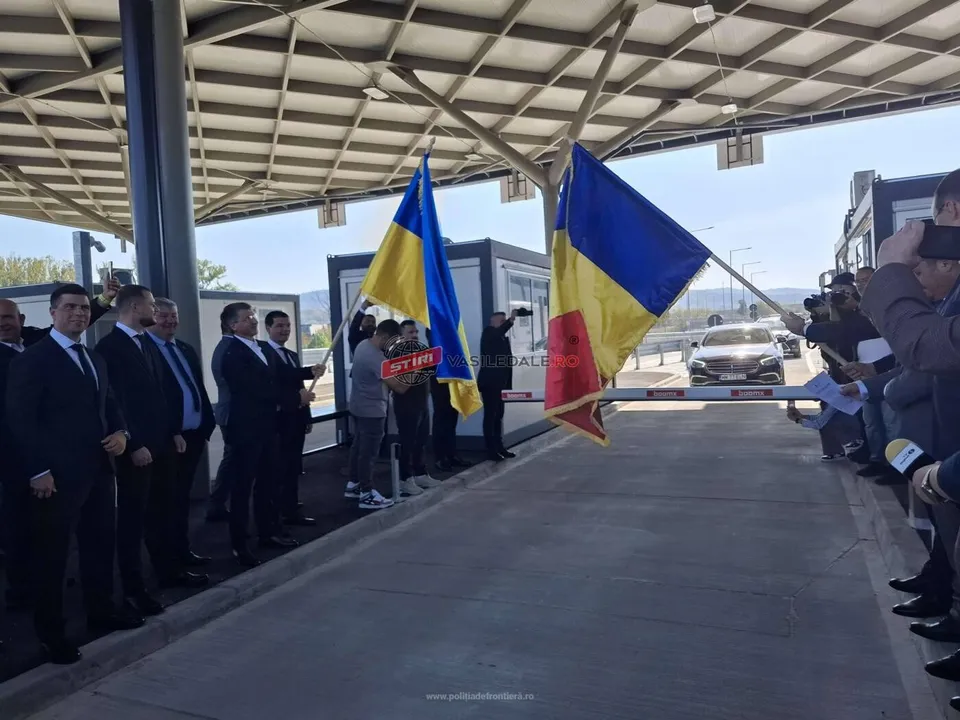 Oamenii stau sub o structură acoperită, purtând steaguri de culoare albastră și galbenă lângă o barieră de culoare albă și roșie.