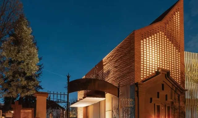O clădire din cărămidă cu fațadă perforată este iluminată sub un cer de seară, lângă un copac înalt și un gard metalic negru.