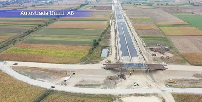 O vedere aeriană prezintă un drum de beton cu două benzi paralele care traversează câmpuri agricole și un pod de beton deasupra unui drum de acces.