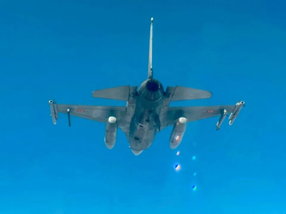 Un avion de luptă gri zboară pe un cer albastru, având sub aripi rezervoare de combustibil și rachete.