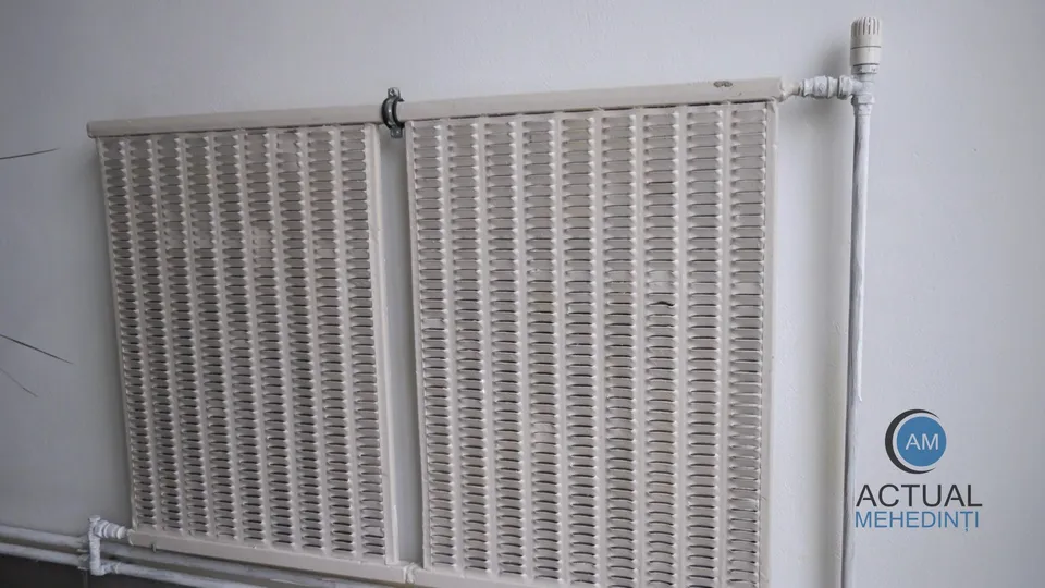 Două radiatoare albe sunt montate pe un perete alb lângă o conductă verticală și o conductă orizontală.