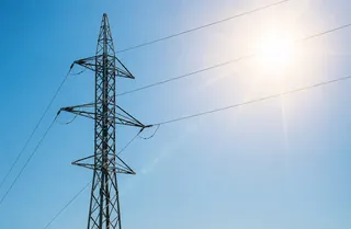 Un stâlp de înaltă tensiune metalic cu cabluri electrice sub un cer albastru luminos cu soare puternic.