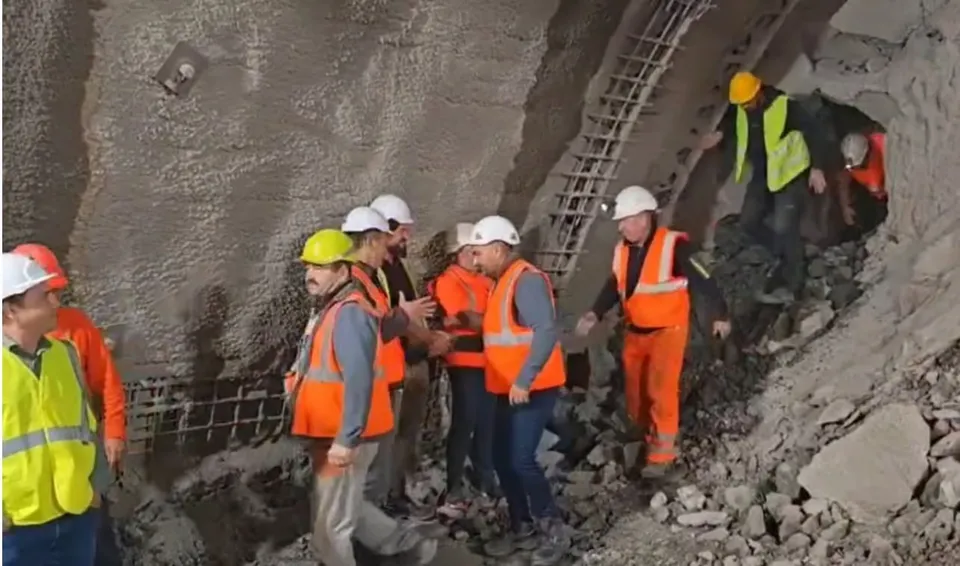 Un grup de lucrători cu căști de protecție și veste reflectorizante se află într-o incintă de tunel cu pereți de beton și resturi de piatră.