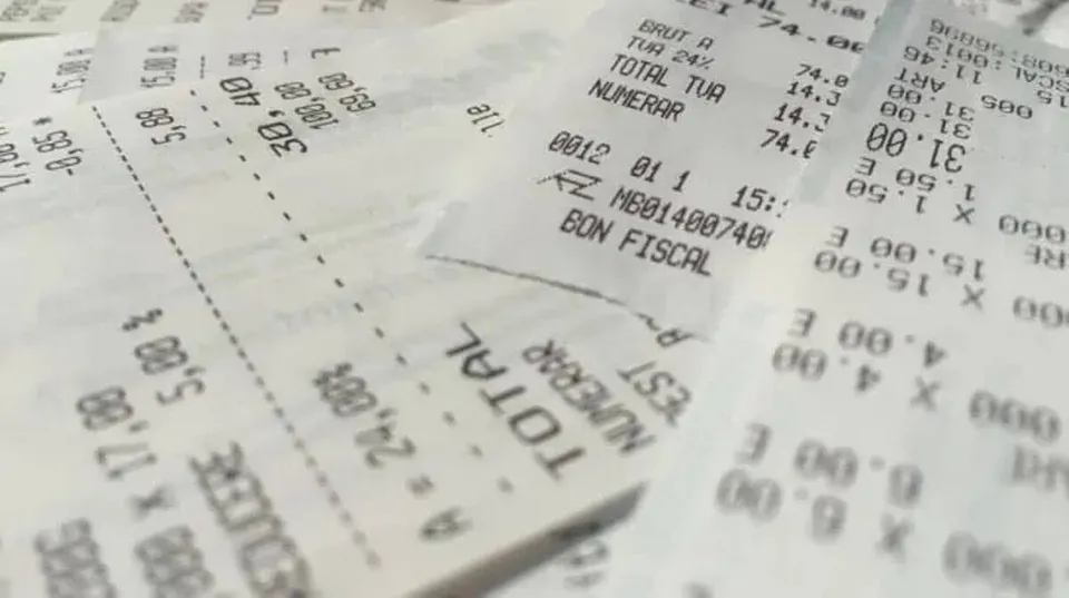 Mai multe bonuri fiscale de hârtie albe sunt împrăștiate pe o suprafață, afișând cifre, text și linii de separare negre.